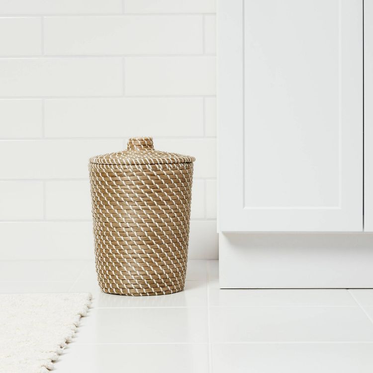 A beige and white colored woven wastebasket with a lid is placed on a white tiled floor next to a cabinet.