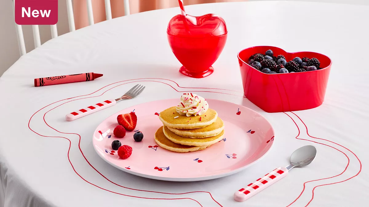 New
A pink plate with a cherry pattern holds pancakes. Red & pink plaid silverware is set out with a heart-shaped bowl full of berries. A red crayon was used to draw a place setting on the white tablecloth & sits to the side. This scene is playful & youthful. 