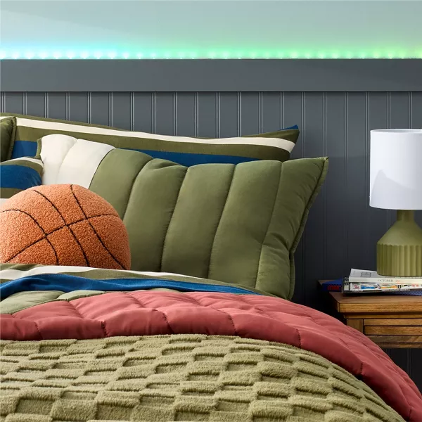 A color block patterned bed spread with deep greens, blues & white, is the focus. A fuzzy football & basketball shaped pillow rest on top of the bed. A mix of textures makes this space cozy, yet the dark colors bring out a masculine edge. 