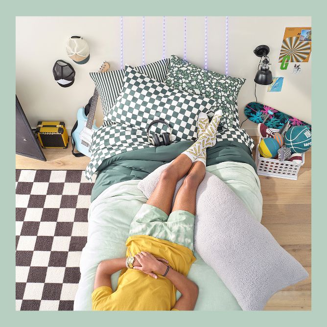 Student laying upside down on their bed in the dorm room, with feet up by checkered pillows and bedsheets.