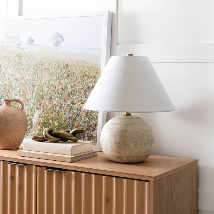 A table lamp with a white colored shade and round base sits on a wooden cabinet next to a vase, a decorative chain, and a framed landscape artwork.