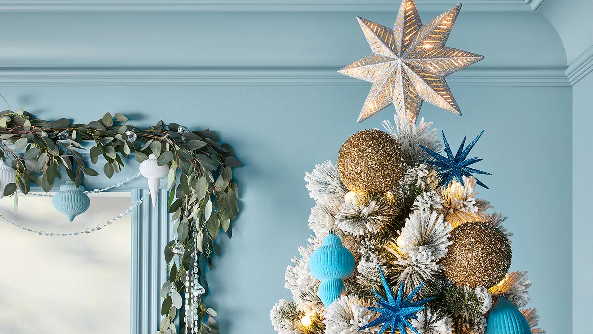 The image shows the top part of a Christmas tree with white-tipped branches. The tree is decorated with large blue and gold-colored ornaments, blue stars, and a bright star on top. Nearby, a garland with green leaves and white ornaments drapes over a window. The walls are light blue.