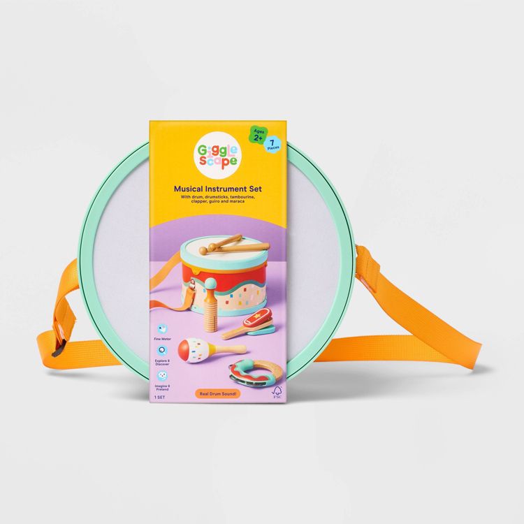 A colorful musical instrument set package featuring a drum, drumsticks, tambourine, clapper, guiro, and maraca, with an orange strap visible.