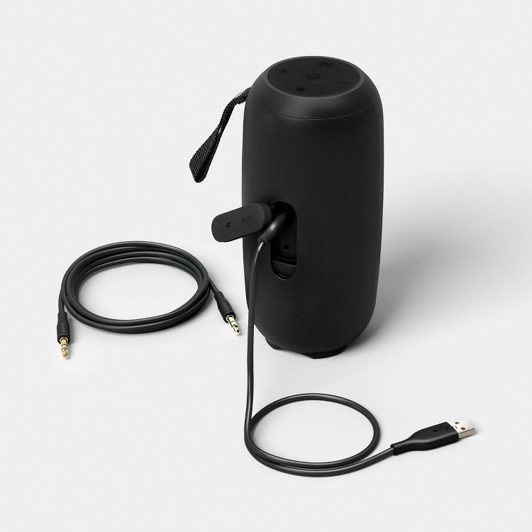 Black colored cylindrical speaker with control buttons on top, an AUX port, and a USB cable connected, alongside a coiled audio cable.