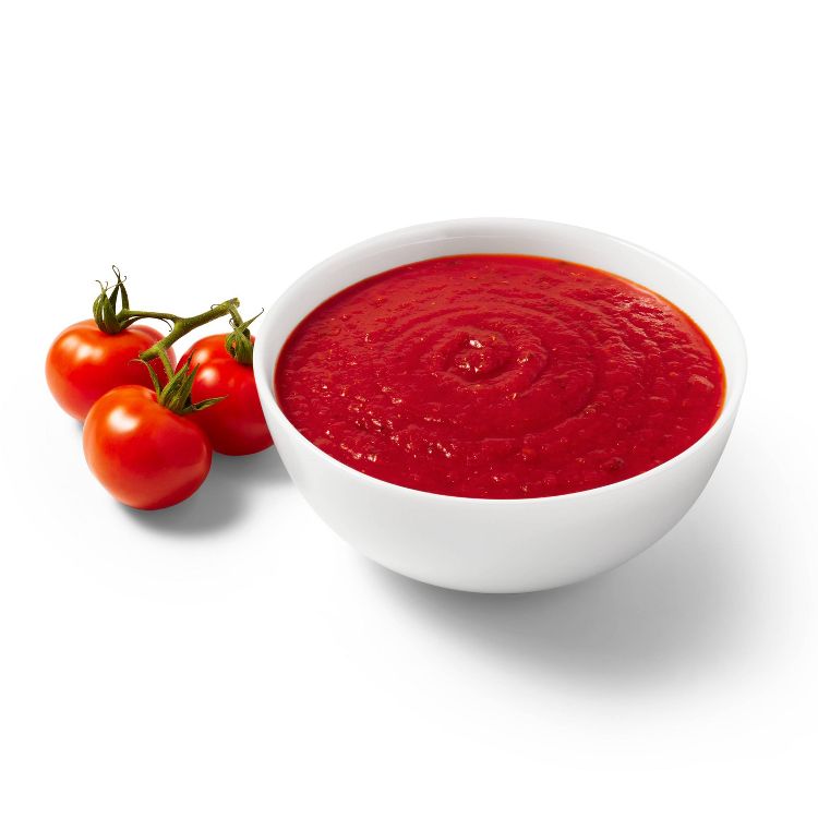 A white bowl filled with red colored tomato sauce next to a cluster of fresh tomatoes.