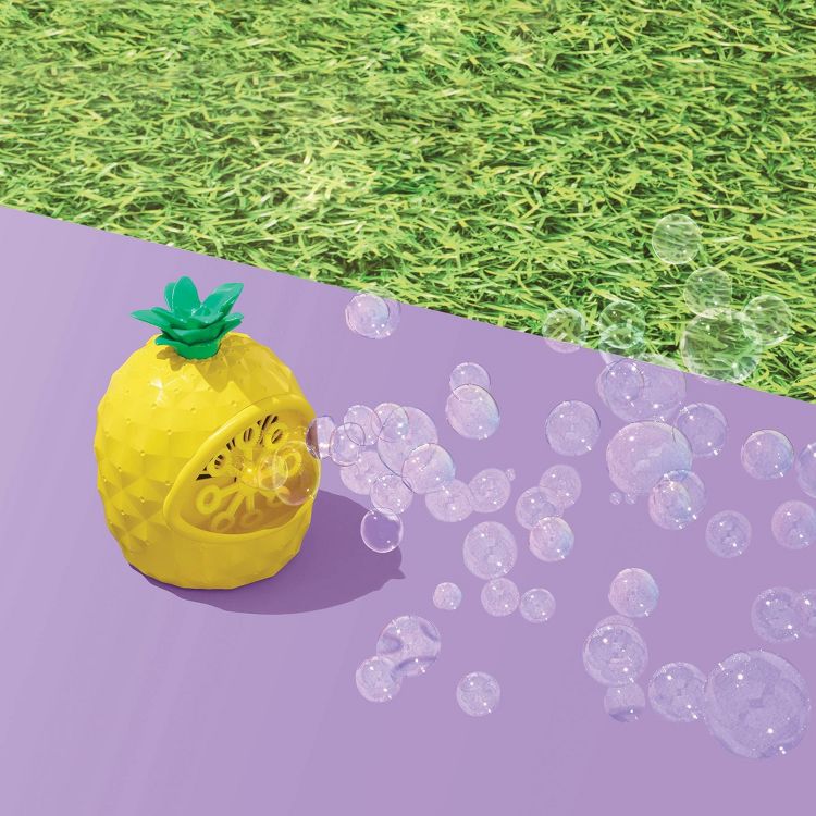 A yellow colored pineapple-shaped bubble blower toy with green leaves, releasing bubbles on a purple surface next to grass.