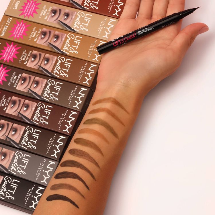 A hand holding a black colored eyebrow pen with multiple brown colored swatches on the arm, next to boxed eyebrow products labeled "Lift & Snatch."