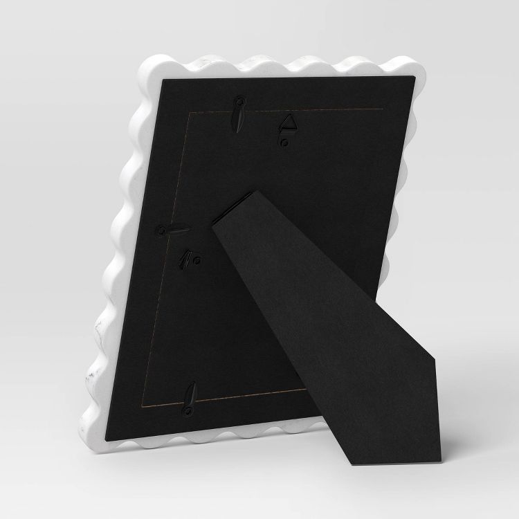 A white colored picture frame with a scalloped edge, shown from the back with a black stand and hanging hooks.