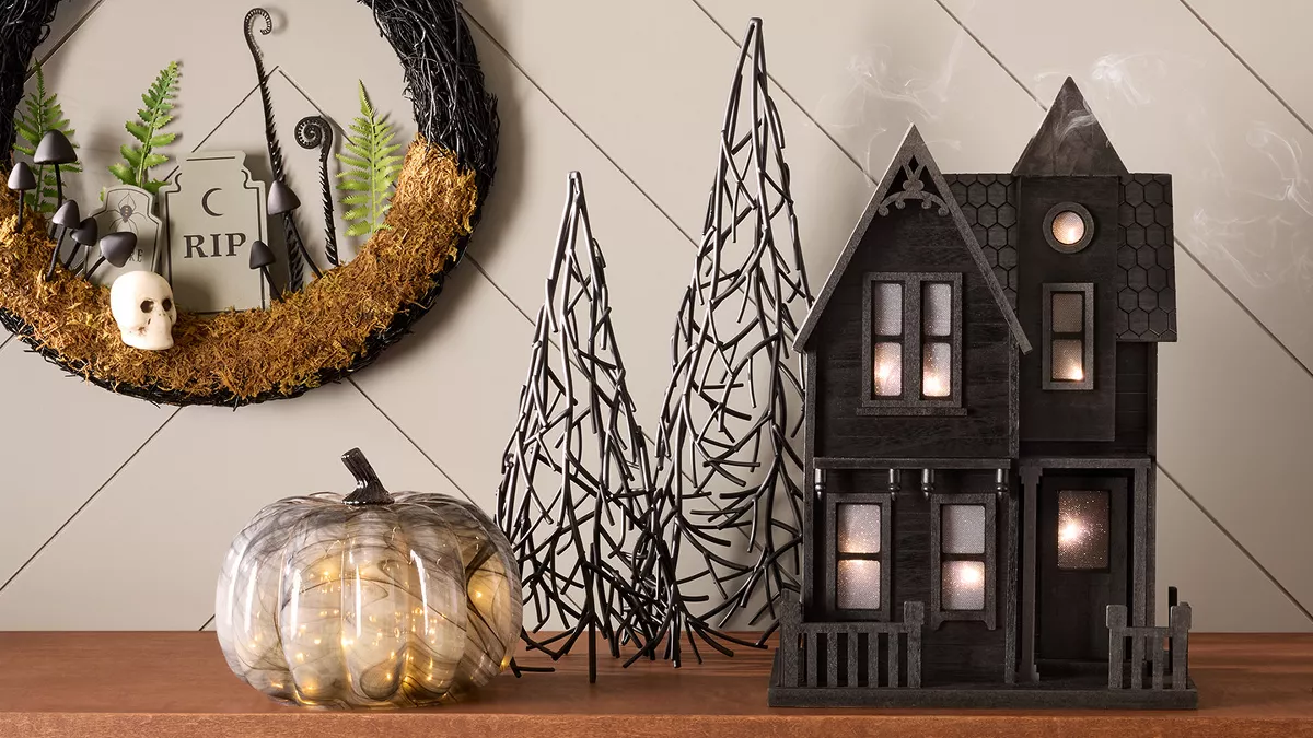 A black 2-story haunted house with lights sits on a table between a gold snake candle holder, 2 black metal trees & a clear glass pumpkin with twinkling lights inside. On the wall, a black twig wreath hangs showing a graveyard scene. 