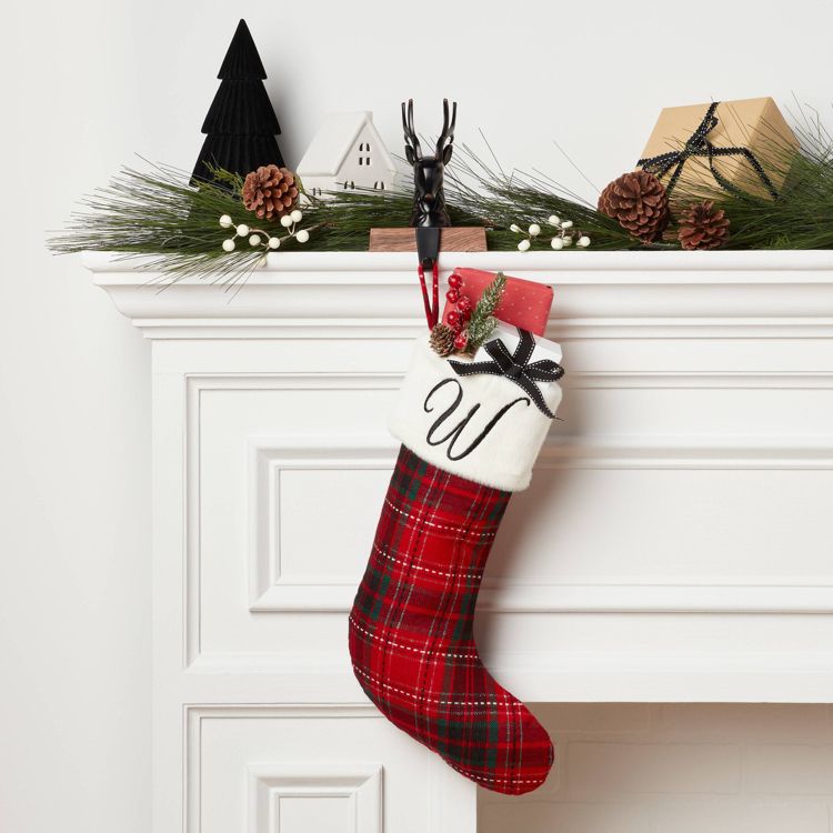 A red and green plaid colored stocking with a white cuff featuring the letter "W" hangs from a mantel decorated with pine branches, pinecones, and small ornaments.