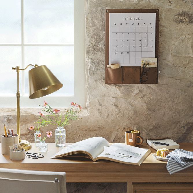 Sunlight streams through the window of an office with a stone wall. A gold lamp, open notebook, and bud vase with pink flowers sits on a wood desk. A calendar with pocket compartments hangs on the back wall.
