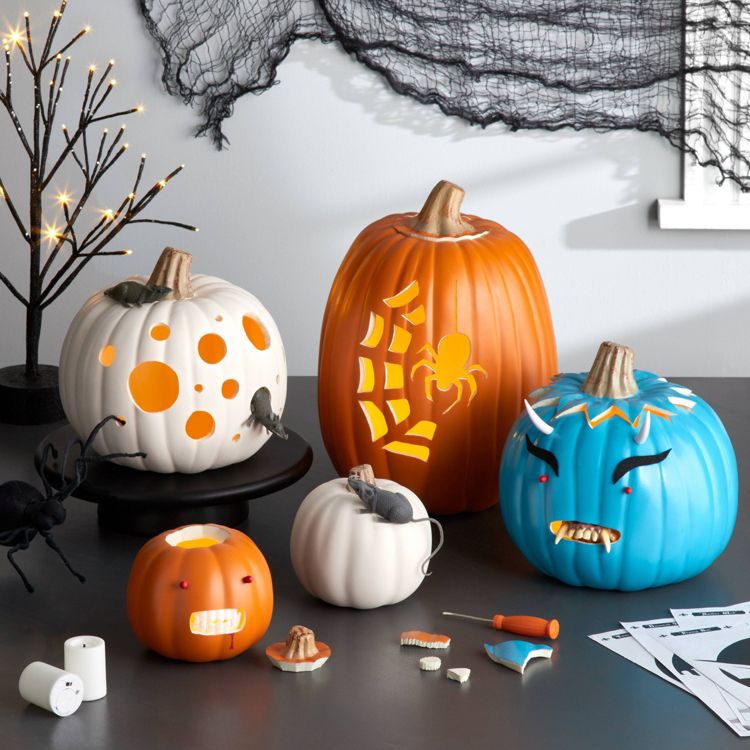 A collection of carved pumpkins in various colors, including orange, white, and blue, with designs featuring a spider, polka dots, and facial expressions, surrounded by Halloween decorations.