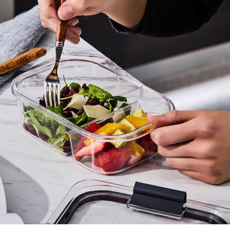A clear rectangular container with two compartments, one holding a salad with greens and cheese, and the other containing sliced strawberries and orange pieces, placed on a marble surface.