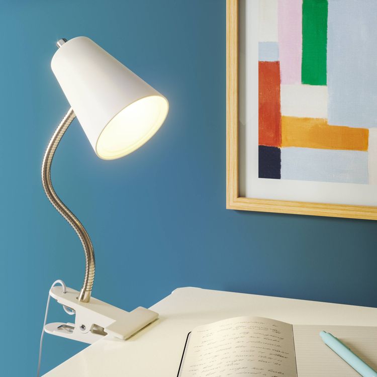 A white colored desk lamp with a flexible neck and clamp base is attached to a table, illuminating an open notebook.