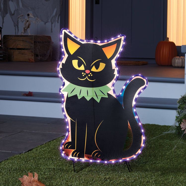 A black cat decoration with a green collar and glowing lights around its outline is placed on a lawn, with pumpkins visible on a porch in the background.