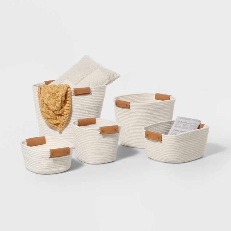 White coiled rope baskets with square bases and tapered sides, featuring brown handles. One basket holds a pillow and a yellow blanket, while another contains a magazine.