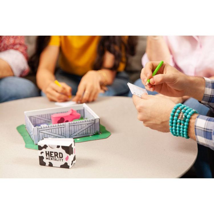 A group of people playing the "Herd Mentality" game with a box and a small pen containing pink colored cow figures on a table.