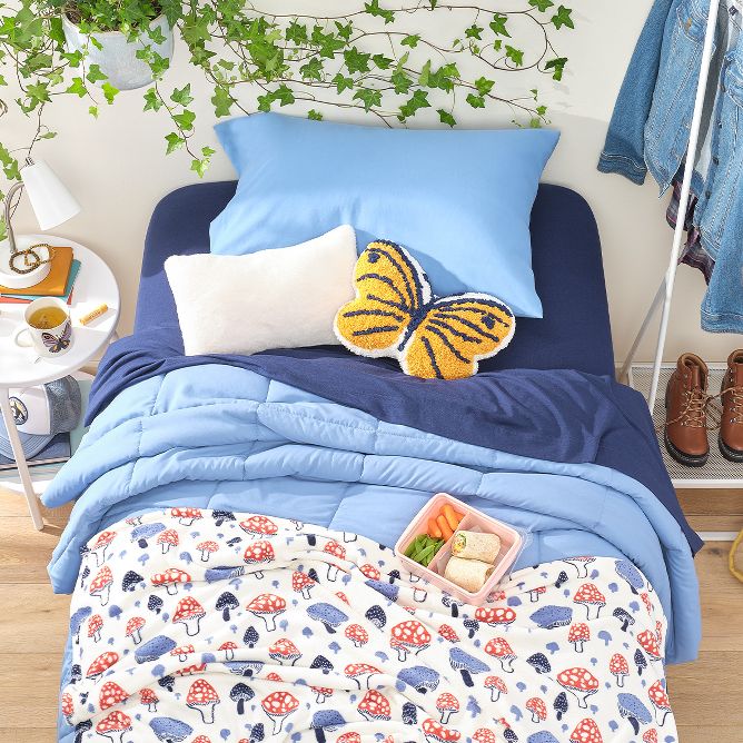 A twin-sized light blue comforter & dark blue sheets center the room, with a mushroom print blanket at the bottom of the bed. On top, a bento box & butterfly throw pillow rest under a growing hanging plant. A clothing rack holds a jean jacket & boots. 