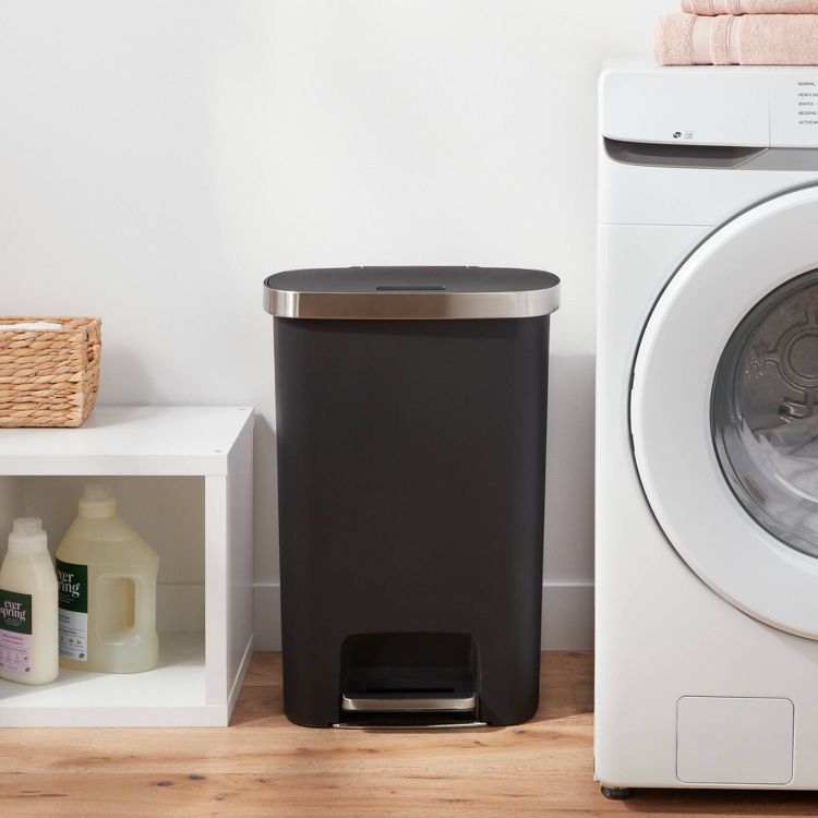 A black colored trash can with a silver lid and foot pedal is placed between a white shelf and a washing machine.