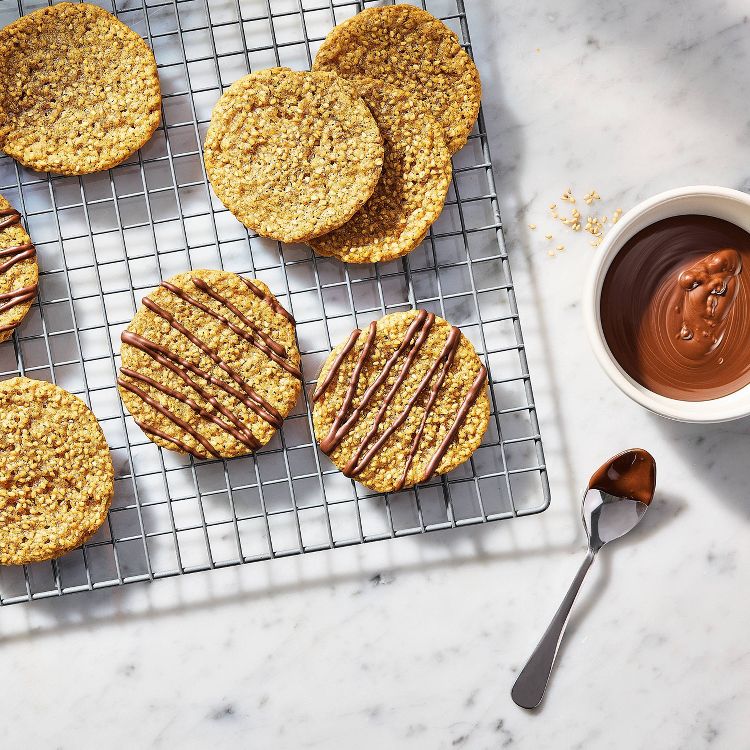 Round cookies with chocolate drizzle on a cooling rack beside a bowl of melted chocolate and a spoon on a marble surface.