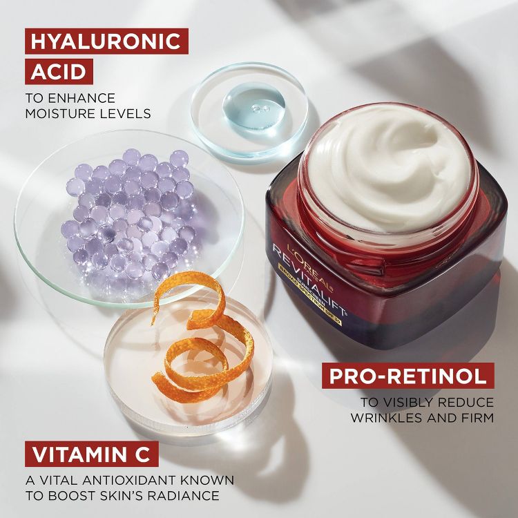 A red jar of L'Oreal Revitalift moisturizer with white cream inside, surrounded by dishes containing blue gel, purple beads, and orange peel, with text highlighting hyaluronic acid, pro-retinol, and vitamin C benefits.