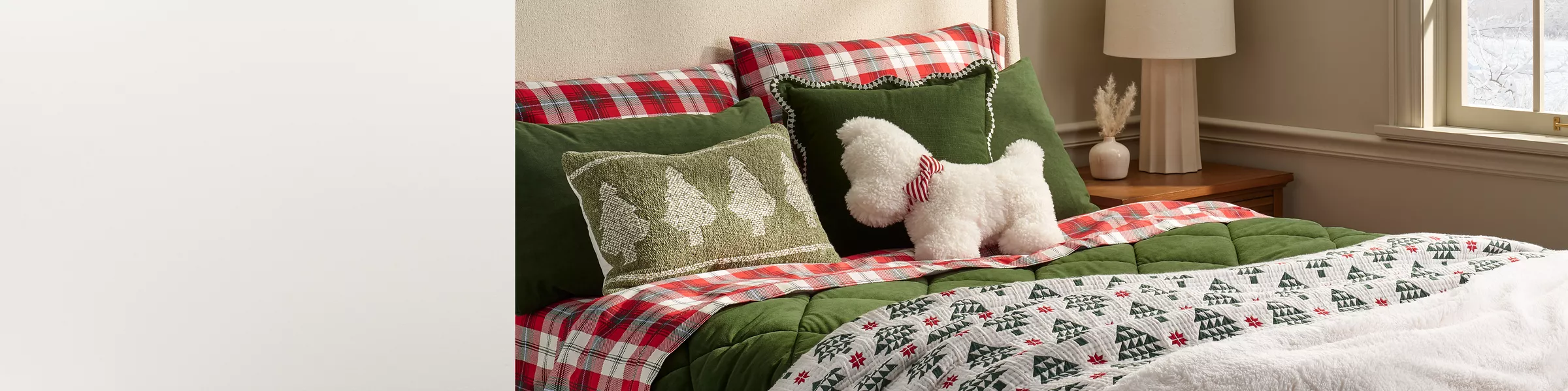 A bed is made with red & green plaid sheets & a green velvet comforter. A Christmas tree patterned quilt lays across the bottom of the bed along with a plush white blanket. A fuzzy white dog pillow with a bowtie rests at the top. It’s very festive & cozy.