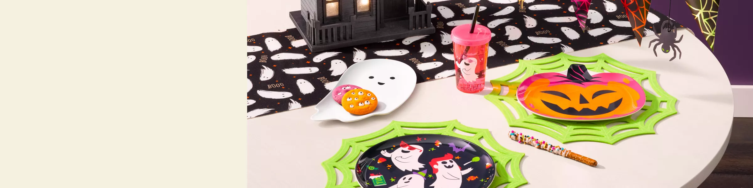 This party decor is kid-friendly & bright. A smiling ghost, pumpkin, & black round plate with 3 ghosts' trick or treating sit on a table holding cookies with eye candies. Green spiderweb placemats are used along with a black table runner with ghosts.