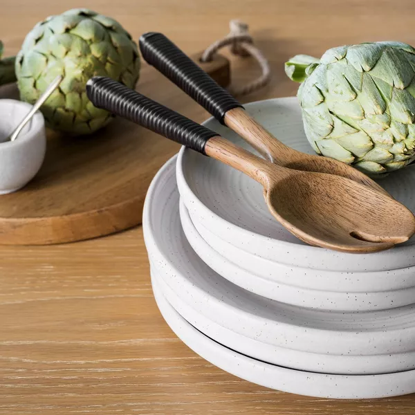 A stack of white plates holding wooden salad servers and artichokes on a wooden table.