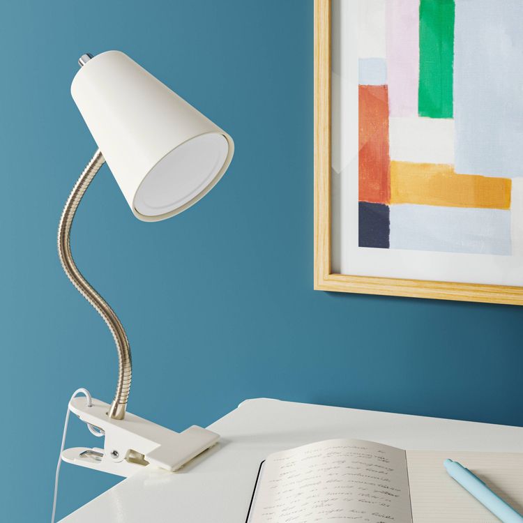 A white colored clip-on desk lamp with a flexible neck is attached to a table beside an open notebook and a framed abstract painting on a blue wall.