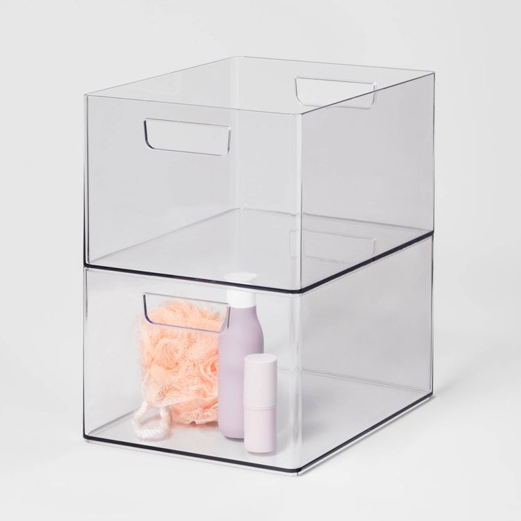 Clear bathroom organizer bin with handles, stacked with another bin. The lower bin contains a pink bottle, a white bottle, and a peach-colored bath sponge.