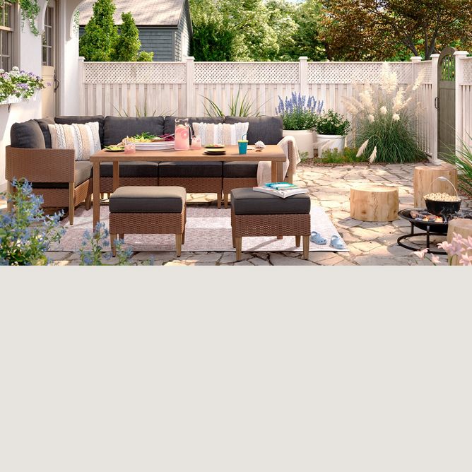 A large rectangular table with a tray, plates & pitcher is centered by a wicker couch & two ottomans. Nearby, a small fire pit pops a bowl of popcorn with wood stumps around it. Various plants bloom along the edges of the patio inside a tall fence. 