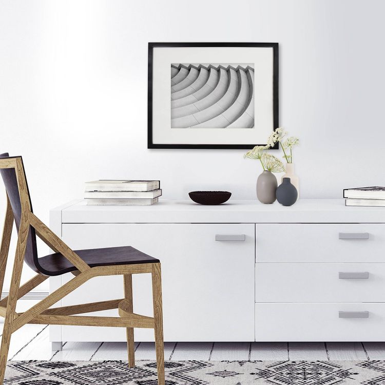 A black-framed monochrome abstract art piece hangs above a white dresser with books, vases, and a bowl, next to a wooden chair with a black seat.