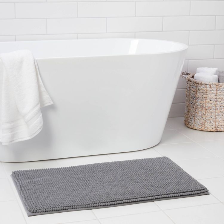 A gray colored bath mat is placed on a white tiled floor in front of a white bathtub with a towel draped over the edge, next to a woven basket.