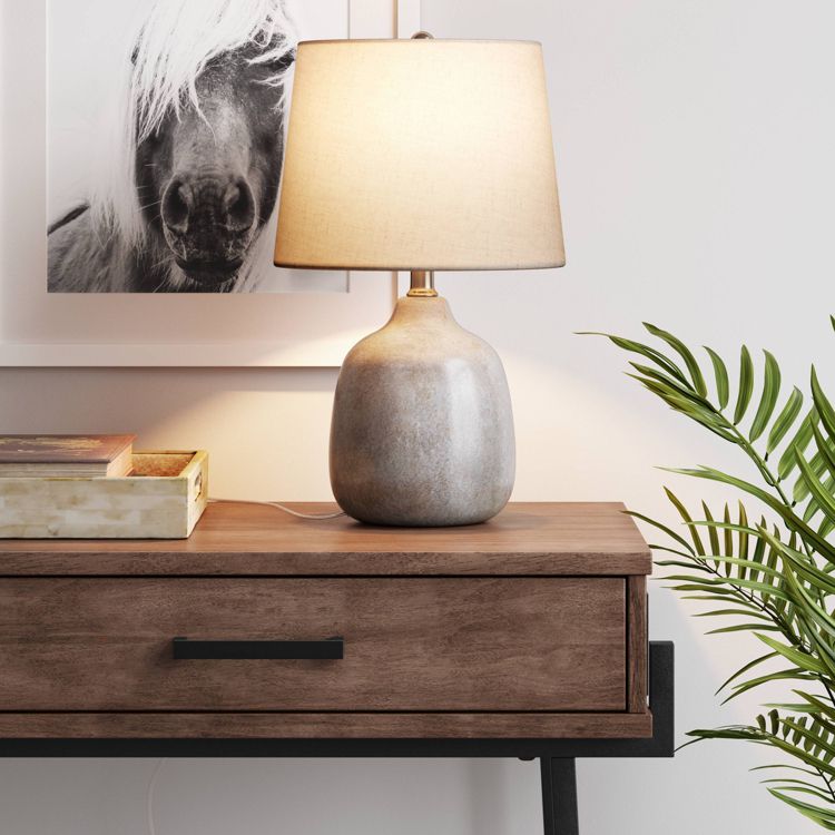A gray colored table lamp with a beige colored shade sits on a wooden table next to a framed black and white photo and a green plant.