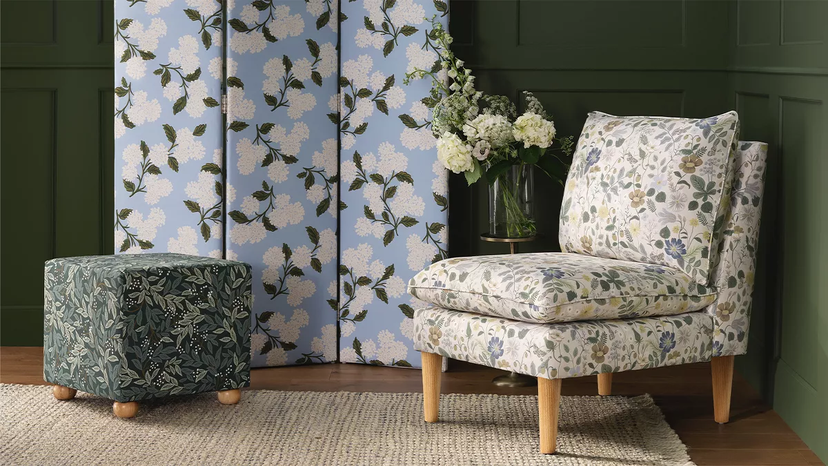 In the corner of a room with deep green walls there’s a white floral accent chair. A light blue room divider with 3 sections & a white hydrangea print stands in front of a wall. There’s a dark green & black ottoman nearby with a leafy design.  