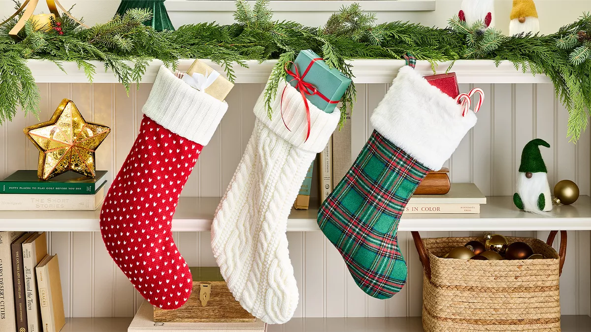 A cozy holiday scene with three stockings hung from a shelf, filled with gifts. The shelf is decorated with green garland, small gnome figures, and Christmas trees in wood-finish and green. Below are books, a gold-colored star, and a basket holding ornaments.