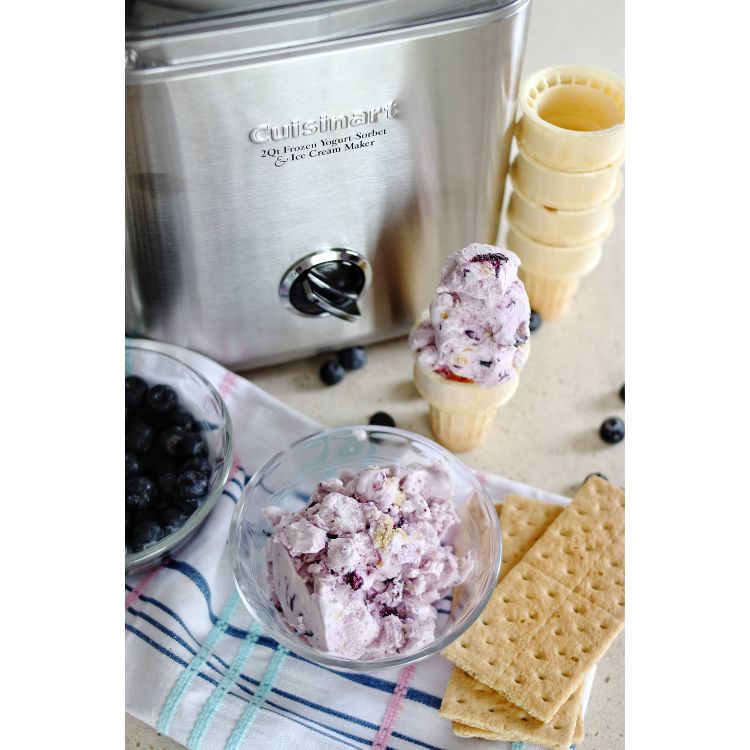 A stainless steel ice cream maker with a bowl of blueberry ice cream, a stack of cones, blueberries, and graham crackers nearby.