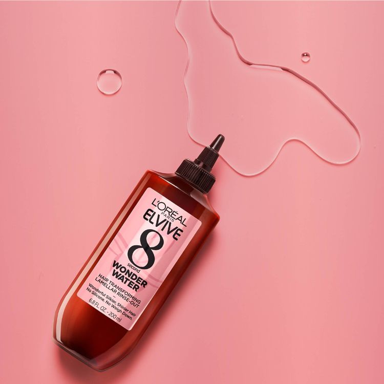 A bottle of L'Oreal Paris Elvive 8 Second Wonder Water Hair Transforming Lamellar Rinse Out with 6.8 FL OZ - 200 ml label on a pink surface with liquid spill.