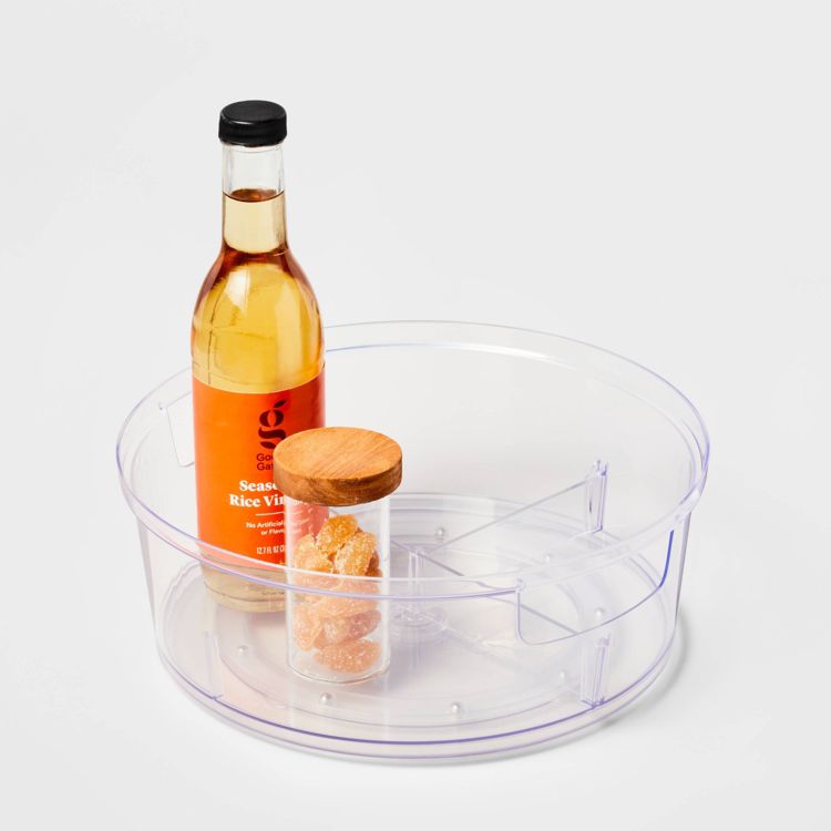 A clear round kitchen storage container holds a bottle of seasoned rice vinegar and a small jar with a wooden lid.