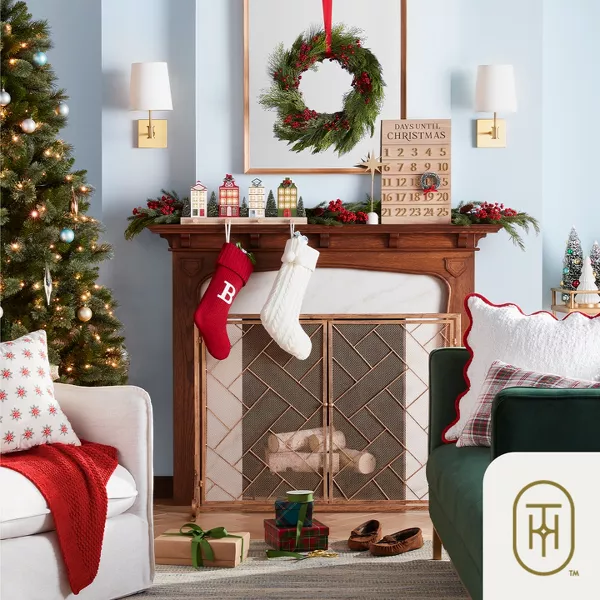 Classic Christmas colors like red & green fill a cozy living room. Throw pillows in various holiday patterns, colors & textures adorn the couch & chair. Tree figures, little house stocking-holders & a Christmas countdown round out the festive scene.