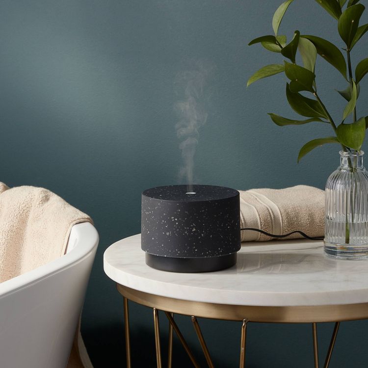 A black colored aromatherapy diffuser emitting mist is placed on a round marble table next to a clear glass vase with green leaves and a beige towel.