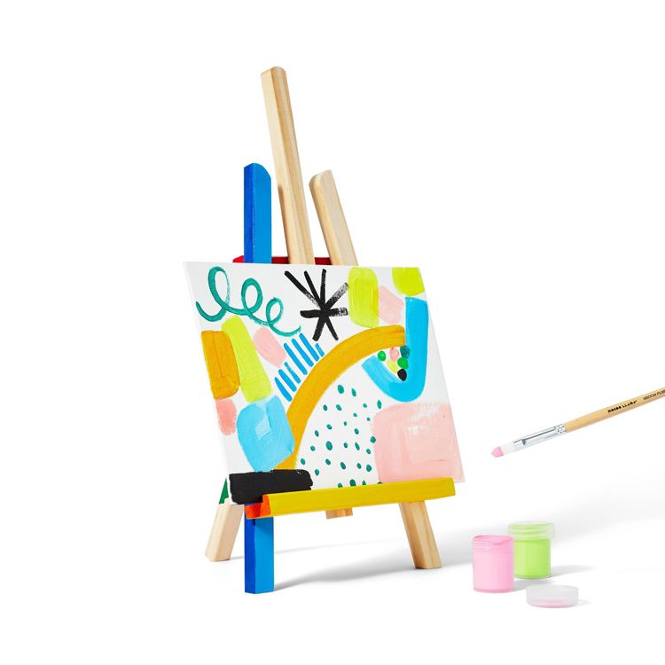 A small wooden easel holds a colorful abstract painting, with a paintbrush and three small jars of pink and green colored paint nearby.